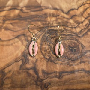 Puede incluir: Un par de pendientes colgantes con pequeños dijes de conchas cauri esmaltadas en color rosa, suspendidos de ganchos de metal dorado. Las conchas tienen un acabado liso y brillante con una abertura vertical central que muestra un interior dorado.