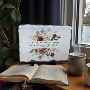 May include: A rectangular decorative slate with a floral design in burgundy, cream, and yellow. The text reads "As for me and my house we will serve the Lord Joshua 24:15". The slate is propped up on black stands, with an open book and a mug of coffee nearby.