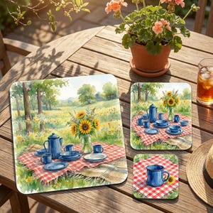May include: A set of three square placemats and coasters featuring a watercolor illustration of a picnic scene. The artwork depicts a red and white checkered blanket on a grassy field with a blue teapot, blue mugs, blue plates, and sunflowers.