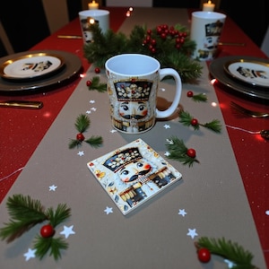 May include: A white ceramic mug and square coaster featuring a Nutcracker design with a detailed face, hat, and uniform. The mug and coaster are on a table with festive decorations, including greenery, red berries, and star-shaped confetti.