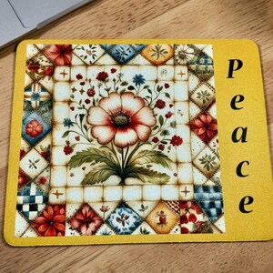May include: A yellow rectangular mousepad with the word "peace" in black vertical text. The center features a floral design with a large red and white flower, surrounded by smaller flowers and green leaves. The border has a patchwork pattern.