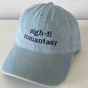 May include: Light blue denim baseball cap with the embroidered text "sigh-fi romantasy" in navy blue. The cap has a curved brim and an adjustable strap at the back. A casual accessory for everyday wear.