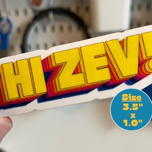 Może przedstawiać: Prostokątna naklejka z ż&oacute;łtym napisem "HI ZEV!" obramowanym tęczowym gradientem. Naklejka ma wymiary 8,9 cm na 2,5 cm. Naklejka ma efekt 3D z białym obramowaniem.