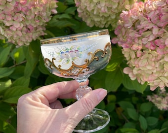 May include: A clear glass cordial glass with a gold rim and ornate gold filigree design. The glass features a white frosted band with painted floral details. The stem is clear and has a decorative, faceted base.