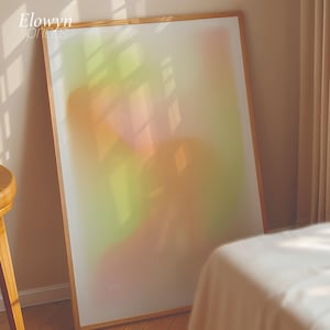 Op de afbeelding: Een ingelijste kunstdruk met een zacht, abstract ontwerp. Het kunstwerk toont gemengde kleuren groen, oranje en roze. De lijst is gemaakt van lichtgekleurd hout. De tekst "Elowyn Prints" staat in de linkerbovenhoek.