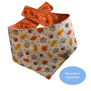 May include: A reversible pet bandana featuring a cream background with a pattern of orange and yellow autumn leaves, brown acorns, and paw prints. The reverse side is orange with white polka dots and includes orange plastic snap buttons for closure.
