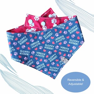 May include: Reversible and adjustable dog bandana with a navy blue side featuring pink paw prints, hearts, and the text "SLOPPY KISSER!". The reverse side is red with white paw prints. Includes a tag.