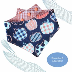 Puede incluir: Bandana reversible y ajustable para mascotas con fondo azul marino y estampado de calabazas naranjas. El reverso presenta un estampado de cuadros. La bandana se asegura con botones naranjas. El texto "Reversible & Adjustable!" está en un círculo azul.