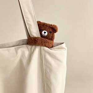 May include: A beige tote bag with a brown teddy bear peeking out from the top. The bag has a long strap and a simple design. The teddy bear has a round head, small ears, and a black nose and eyes.