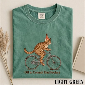 May include: A light green t-shirt featuring a graphic of a brown tabby cat wearing a red party hat with yellow polka dots, riding a bicycle. Below the cat, the text reads "Off to Commit Dad Foolery" in a dark brown, sans-serif font.