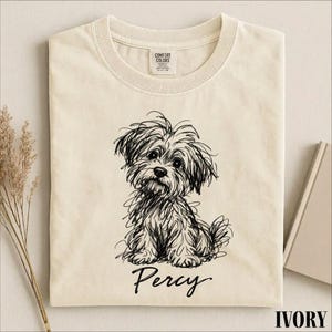 May include: An ivory-colored t-shirt featuring a black line-art illustration of a shaggy dog sitting down. Below the dog, the name Percy is printed in a black cursive script. The shirt has a crew neckline and a Comfort Colors brand label.