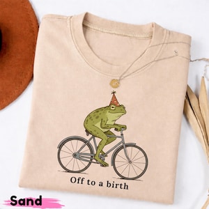 May include: A sand-colored t-shirt featuring a graphic of a green frog wearing a red and yellow polka-dot party hat while riding a bicycle. Below the illustration, the text reads "Off to a birth" in a simple black sans-serif font.