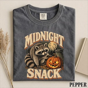 May include: A charcoal gray crew neck t-shirt featuring a graphic of a raccoon next to a jack-o'-lantern pumpkin. The text MIDNIGHT SNACK is printed in a distressed font above and below the raccoon. The shirt has a Comfort Colors brand label.
