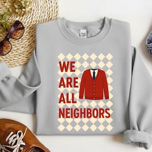 Könnte beinhalten: Ein hellgraues Sweatshirt mit dem Schriftzug "WE ARE ALL NEIGHBORS" in roten Blockbuchstaben. Ein roter Cardigan mit schwarzer Krawatte befindet sich in der Mitte des Designs. Der Hintergrund ist ein Muster aus abwechselnd weißen und hellgrauen Rauten.