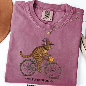 May include: A berry-colored short-sleeve t-shirt featuring a graphic of a brown tabby cat wearing a purple witch hat and riding a bicycle. The cat carries a small orange jack-o'-lantern. The text reads OFF TO BE SPOOKY. The label says COMFORT COLORS.
