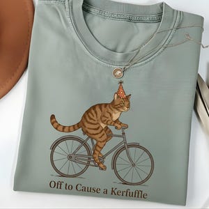 Puede incluir: Camiseta verde salvia con un gato atigrado marrón que lleva un gorro de fiesta y monta en bicicleta. El texto "Off to Cause a Kerfuffle" está impreso debajo de la ilustración. La camiseta tiene cuello redondo y mangas cortas.