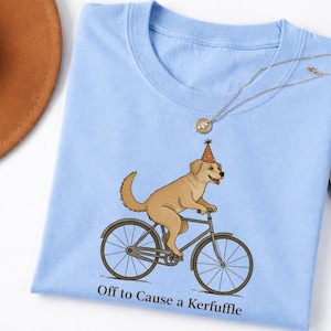 Könnte beinhalten: Hellblaues T-Shirt mit einem Cartoon-Hund auf einem Fahrrad, der einen Partyhut trägt. Der Text "Off to Cause a Kerfuffle" ist darunter aufgedruckt. Eine goldene Halskette liegt auf dem T-Shirt.
