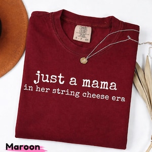 Puede incluir: Camiseta granate con el texto blanco "just a mama in her string cheese era". Un collar plateado con un colgante descansa sobre la camiseta. La camiseta tiene una etiqueta Comfort Colors.