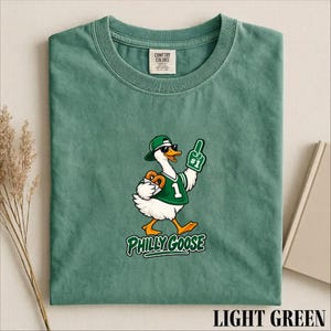 Könnte beinhalten: Ein hellgrünes T-Shirt mit einer Grafik einer weißen Gans, die eine grüne Mütze, eine Sonnenbrille und ein grünes Trikot mit der Nummer 1 trägt. Die Gans hält eine Brezel und einen Schaumstofffinger mit der Aufschrift #1. Darunter steht PHILLY GOOSE.