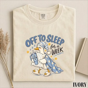 May include: An ivory t-shirt featuring a graphic of a duck wearing a blue and white striped nightcap, holding a pillow and a blue tumbler labeled DO NOT DISTURB. The duck is wrapped in a blue blanket with white daisies and the text OFF TO SLEEP for a WEEK.