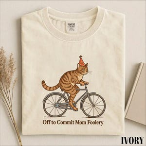Op de afbeelding: Een ivoorkleurig t-shirt met een afbeelding van een bruine cyperse kat die een rood met geel gestippelde feesthoed draagt terwijl deze op een fiets rijdt. Onder de kat staat de tekst, Off to Commit Mom Foolery. Het shirt heeft een ronde hals.