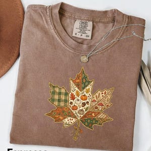 Op de afbeelding: Een bruin t-shirt met korte mouwen en een grote esdoornblad-afbeelding verdeeld in secties met patronen van pompoenen, paddenstoelen, eikels en ruitjes. Het binnenlabel toont Comfort Colors S P CH. Het woord Espresso staat eronder gedrukt.