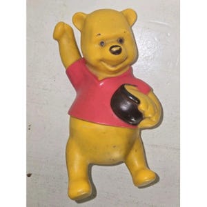 May include: A yellow Winnie the Pooh figurine with a red shirt and a black honey pot. The figurine is waving with one arm raised. The toy is made of plastic and has a vintage appearance.