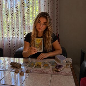 May include: A tarot card reading setup featuring a deck of cards, with one card held up displaying The Sun, alongside a bundle of dried sage, three small wooden runes, a raw amethyst crystal, and a clear glass sphere on a gold-toned stand.