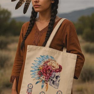 Könnte beinhalten: Beige Tragetasche mit einem Totenkopf, der einen Blumenschmuck mit blauen und roten Federn trägt. Die Tasche hat auch drei Traumfänger-Designs. Die Person auf dem Bild trägt ein braunes Kleid und ein Federstirnband.