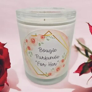 Peut inclure: Une bougie parfumée blanche dans un pot en verre avec un couvercle transparent. L'étiquette indique "Bougie Parfumée 'For Her'" avec des éléments floraux et géométriques. La bougie est posée sur une surface rose, avec une rose rouge en arrière-plan.