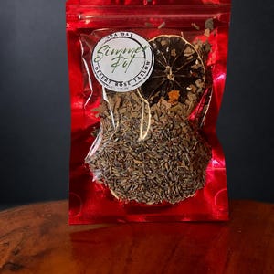 May include: A shiny red plastic pouch containing a mixture of dried lavender buds, wood chips, and a single dried citrus slice. A circular white label on the front features the text "SPA DAY", "Simmer Pot", and "DESERT ROSE TALLOW" in black ink.