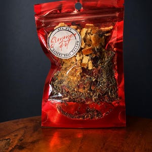 May include: A shiny red resealable pouch containing a dried botanical blend of orange peels, lavender, and flower petals. A circular white label on the front features the text Simmer Pot, Spa Day, and Desert Rose Tallow in black and red ink.