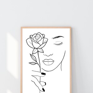May include: Black and white line art print of a woman's face holding a rose. The artwork features a minimalist style with closed eyes and a detailed rose. The print is framed in a light brown wooden frame.