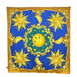 May include: A square silk scarf with a celestial design. The scarf features a blue center with gold suns, crescent moons, and stars. A gold border frames the design. The scarf has the word "LANCEL" printed on the bottom right corner.
