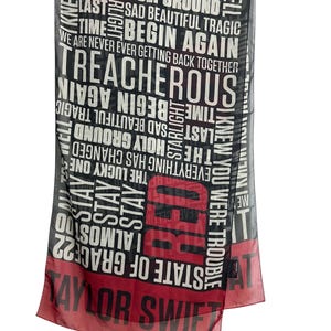 May include: A rectangular scarf with a black background and red accents. The scarf features the word "RED" in large red letters, along with song titles and the name "Taylor Swift" in white. The scarf is made of a sheer material.