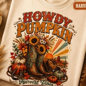 May include: A cream-coloured t-shirt featuring a graphic print with the text "HOWDY PUMPKIN" and "Harvest Vibes". The design includes cowboy boots filled with sunflowers, pumpkins, a horseshoe, a cowboy hat, and a desert sunset background.