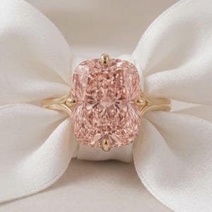 Puede incluir: Un anillo de oro con una gran piedra preciosa rectangular de color melocotón en talla cojín, sujeta por cuatro garras. La banda es lisa y pulida, mostrando un diseño clásico que resalta la piedra facetada y su tono cálido y suave.