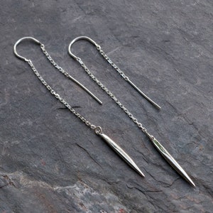 May include: A pair of silver dangle earrings with a long chain and a pointed, silver pendant. The earrings are made of sterling silver and are perfect for adding a touch of elegance to any outfit.