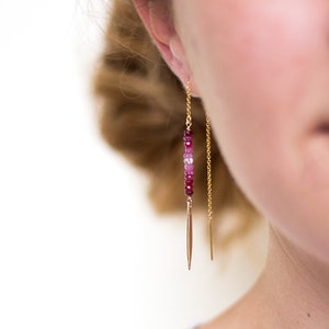 May include: Gold chain earrings with a pink and red gemstone dangle. The earrings have a pointed tip at the end of the chain.