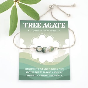 May include: A silver chain bracelet with three green and white tree agate beads. The bracelet has a sliding clasp and is displayed on a card that reads "TREE AGATE + Crystal of Inner Peace + CONNECTED TO THE HEART CHAKRA, TREE AGATE IS SAID TO PROVIDE A SENSE OF TRANQUILITY & PROMOTE PROSPERITY."
