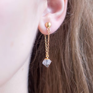 May include: Gold chain earrings with a clear crystal pendant. The earrings are dangling from the earlobe.