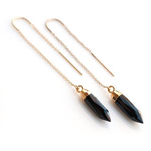 Op de afbeelding: Een paar gouden kettingoorbellen met zwarte edelsteen druppel hangers. De edelstenen zijn gefacetteerd en hebben een gouden metalen setting.