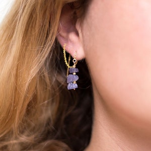 May include: A pair of gold earrings with a chain connecting them. The earrings feature a cluster of three purple amethyst stones.