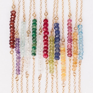 May include: A set of 12 delicate gold chain necklaces, each featuring a single row of colorful gemstone beads. The necklaces are arranged in a rainbow pattern, with each color representing a different gemstone. The gemstones include amethyst, aquamarine, emerald, ruby, peridot, sapphire, opal, citrine, topaz, turquoise, and garnet.