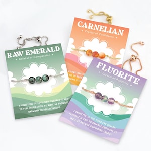 May include: Three gemstone bracelets on cards. The cards are labeled "Raw Emerald", "Carnelian", and "Fluorite". Each bracelet features three beads. The "Raw Emerald" bracelet has dark green beads, the "Carnelian" bracelet has orange beads, and the "Fluorite" bracelet has purple beads.