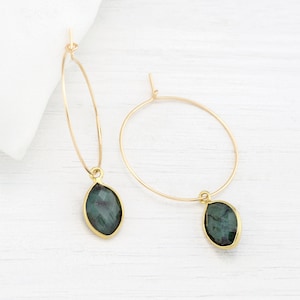 May include: Gold hoop earrings with a dangling green gemstone pendant. The earrings are simple and elegant.