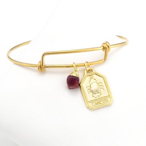 May include: Gold adjustable bangle bracelet with a charm featuring a crab and the word "Cancer". A small, dark red gemstone is also attached to the bracelet.