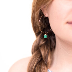 May include: Gold hoop earrings with a green gemstone dangle. The earrings are worn by a person with long brown hair.