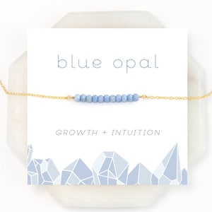 Op de afbeelding: Een delicate gouden ketting met een rij lichtblauwe opaal kralen. De kaart heeft de tekst "blue opal" en "growth + intuition".