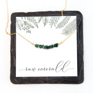 May include: A gold chain necklace featuring a row of small, raw, dark green emerald chips. The necklace is displayed on a white card with green botanical line drawings at the top and the text "raw emerald" in a black cursive script at the bottom.